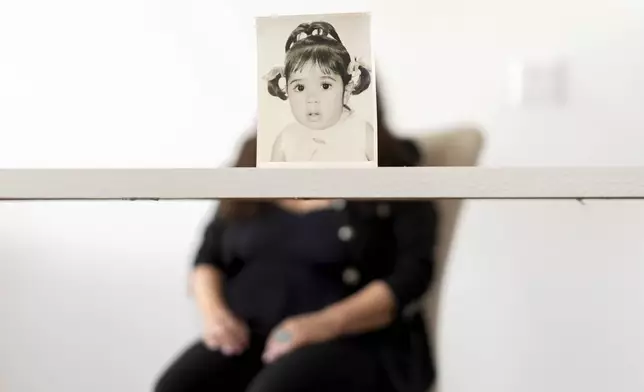 Buttons, who the Associated Press is referring to only by her childhood nickname because of her legal status, sits for a portrait behind her baby photo taken before she was adopted from Iran to a family in America, Monday, June 24, 2024, in Henderson, Nev. Buttons is one of thousands of children adopted from abroad by American parents, many of them military service members, who were left without citizenship by loopholes in American law that Congress has been aware of for decades, yet remains unwilling to fix. (AP Photo/David Goldman)