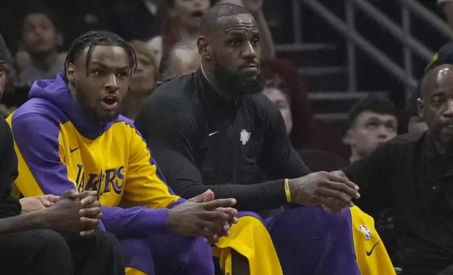Los Angeles Lakers guard Bronny James, left, and forward LeBron James, right, watch from the bench in the first half of an NBA basketball game against the Cleveland Cavaliers, Wednesday, Oct. 30, 2024, in Cleveland. (AP Photo/Sue Ogrocki)