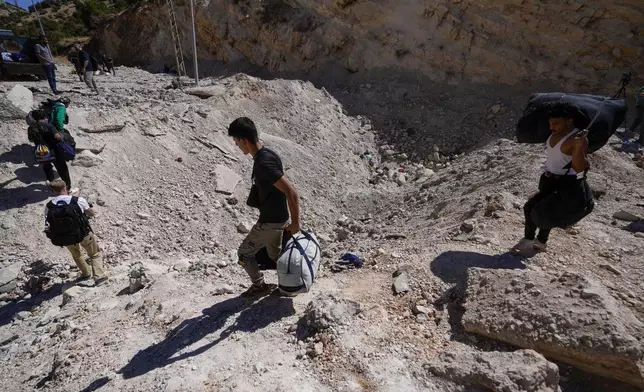 People carry their luggage as they cross into Syria on foot, through a crater caused by Israeli airstrikes aiming to block Beirut-Damascus highway at the Masnaa crossing, in the eastern Bekaa Valley, Lebanon, Saturday, Oct. 5, 2024. (AP Photo/Hassan Ammar)