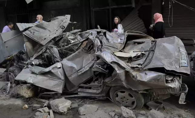 People pass by a destroyed car at the site of Thursday's Israeli airstrike, in Beirut, Lebanon, Friday, Oct. 11, 2024. (AP Photo/Hussein Malla)