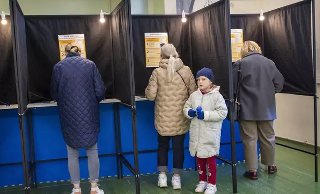 A girl looks from a voting booth as her relative fills-in a ballot at a polling station during the first round of voting in a parliamentary election, in Vilnius, Lithuania, Sunday, Oct. 13, 2024. (AP Photo/Mindaugas Kulbis)