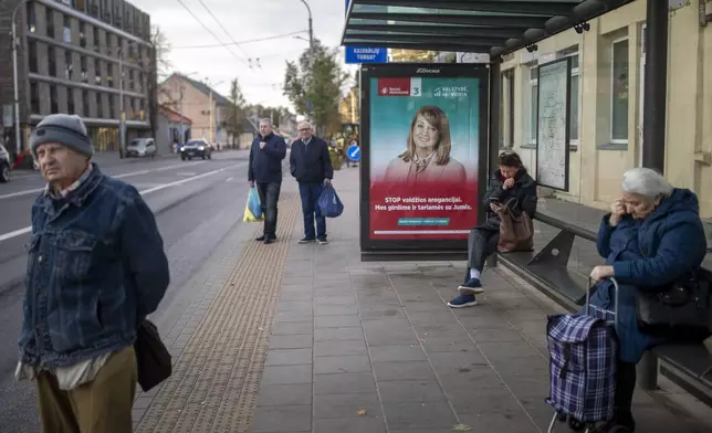 Local residents wait at a bus stop, with a poster displaying leader of the Social Democratic Party Vilija Blinkeviciute in Vilnius, Lithuania, Wednesday, Oct. 9, 2024. (AP Photo/Mindaugas Kulbis)