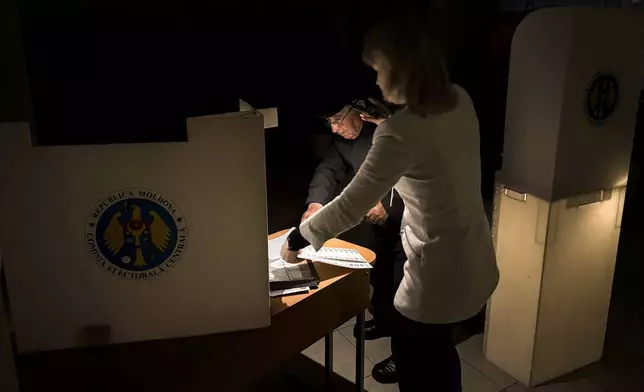 An election official assists a voter with her mobile phone flashlight during a power failure in Chisinau, Moldova, Sunday, Oct. 20, 2024, during a presidential election and a referendum on whether to enshrine in the Constitution the country's path to European Union membership. (AP Photo/Nicolae Dumitrache)