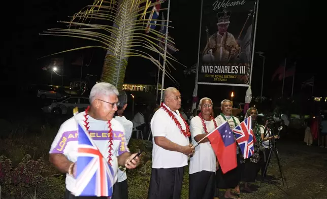 Men hold flags as they wait for the arrival of Britain's King Charles III and Queen Camilla in the village of Siumu, Samoa, on Wednesday, Oct. 23, 2024. (AP Photo/Rick Rycroft)