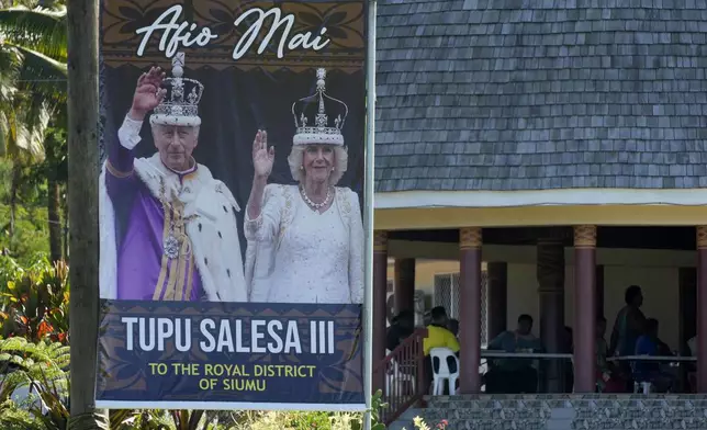 People gather together at a meeting place near a portrait of King Charles III and Queen Camilla in the village of Siumu, Samoa, on Monday, Oct. 21, 2024, as the village prepares for the arrival of the royals. (AP Photo/Rick Rycroft)