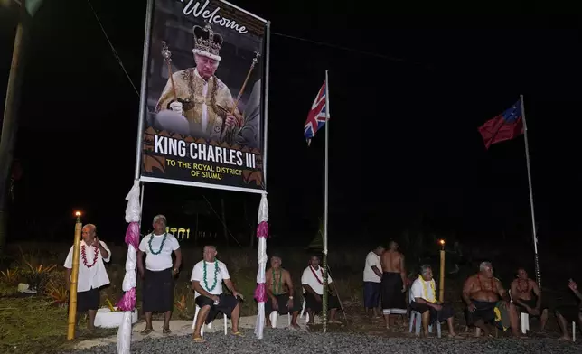 People sit under a portrait of Britain's King Charles III as they wait for his arrival and Queen Camilla in the village of Siumu, Samoa, on Wednesday, Oct. 23, 2024. (AP Photo/Rick Rycroft)