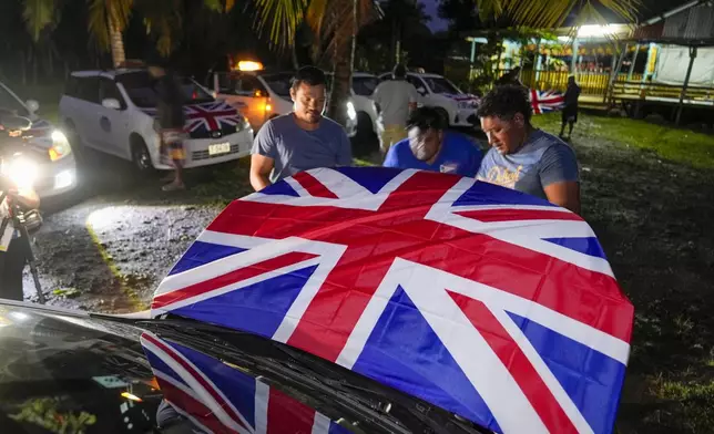 Men attach Union Jacks to the hoods of their taxis as they wait for the arrival of Britain's King Charles III and Queen Camilla in the village of Siumu, Samoa, on Wednesday, Oct. 23, 2024. (AP Photo/Rick Rycroft)