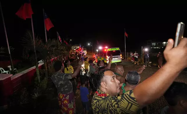 People wave and take pictures as the motorcade with Britain's King Charles III and Queen Camilla passes in the village of Siumu, Samoa, on Wednesday, Oct. 23, 2024. (AP Photo/Rick Rycroft)