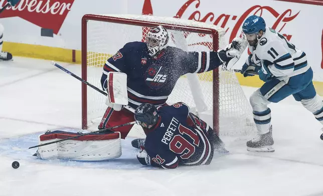 Winnipeg Jets goaltender Connor Hellebuyck (37) saves the shot from San Jose Sharks' Luke Kunin (11) as Cole Perfetti (91) defends during the first period of an NHL hockey game in Winnipeg, Manitoba, Friday, Oct. 18, 2024. (John Woods/The Canadian Press via AP)