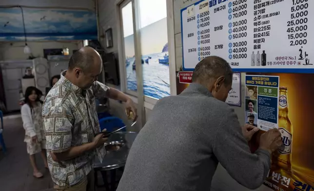 Kenneth Barthel, left, helps Shin Byung-chul post a flyer with photos of Barthel at various ages in his life, on the wall of his restaurant in Busan, South Korea, Friday, May 17, 2024, as Barthel's daughter, Amiya, rear, looks on. Barthel's mother had ordered him soup in a restaurant in the area when he was 6 years old, went to the bathroom and never returned. (AP Photo/Jae C. Hong)