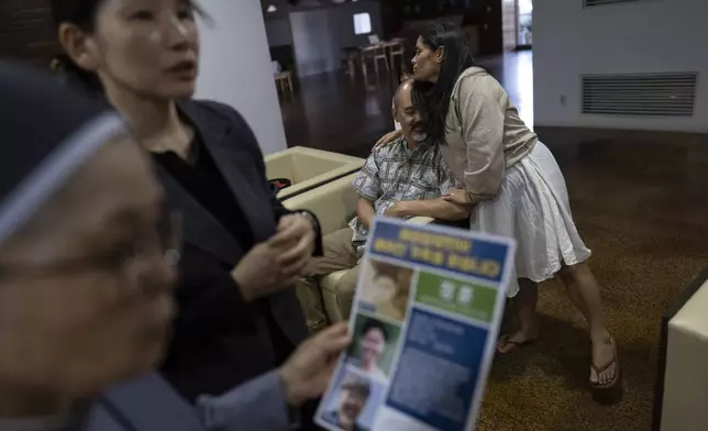 Kenneth Barthel, who was adopted by a single parent in Hawaii at 6 years old, is hugged by his wife, Napela, at the Sisters of Mary in Busan, South Korea, Friday, May 17, 2024. In the foreground, Sister Bulkeia, left, and Paek Kyeong-mi from Global Overseas Adoptees' Link discuss a flyer designed to uncover the details of Barthel's early life and find his birth family. (AP Photo/Jae C. Hong)