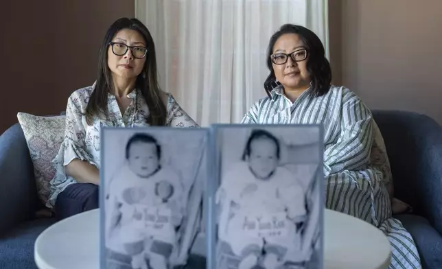 Dee Iraca, left, and her twin sister, Becca Webster, sit behind their baby photos from before they were adopted out of South Korea to a family in the United States, Saturday, April 6, 2024, at Iraca's home in Davidson, N.C. After all, they ended up happy in America. Yet their happiness was built on an injustice that hurt thousands, including their birth father and were too late to meet their mother. (AP Photo/David Goldman)