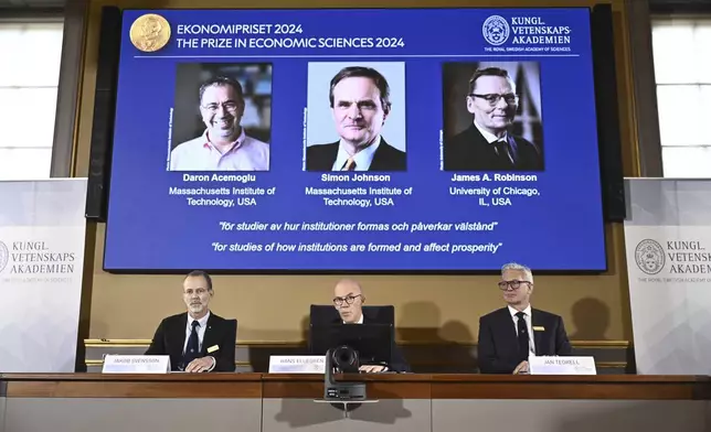 Academy of Sciences permanent secretary Hans Ellegren, center, Jakob Svensson, left, and Jan Teorell, of the Nobel assembly announce the Nobel memorial prize in economics winners, Daron Acemoglu, Simon Johnson and James A Robinson, seen on screen, during a press meeting at the Royal Swedish Academy of Sciences in Stockholm, Sweden, Monday Oct. 14, 2024. (Christine Olsson/TT News Agency via AP)