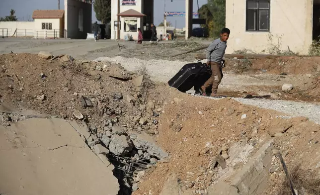 A Syrian boy fleeing the war in Lebanon carries his luggages as he passes through a crater caused by an Israeli airstrike, which blocks the road between the Lebanese and the Syrian crossing points, in Jousieh, Syria, Sunday, Oct. 27, 2024. (AP Photo/Omar Sanadiki)