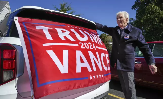 Douglas MacDonald, of Rutherfordton, attaches a flag to the back of his car at the Rutherford County Annex Building where early voting was taking place, Thursday, Oct. 17, 2024 in Rutherfordton, N.C. (AP Photo/Kathy Kmonicek)