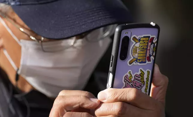 A man uses a mobile phone with a decoration to celebrate an achievement of Shohei Ohtani of the Los Angeles Dodgers as he and others gather to watch a live stream of Game 3 of the baseball World Series between the Dodgers and the New York Yankees during a public viewing event in Oshu, northeastern Japan, the hometown of Shohei Ohtani of the Los Angeles Dodgers, Tuesday, Oct. 29, 2024. (AP Photo/Eugene Hoshiko)