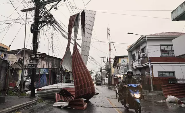 Motorists ride past a part of a roof suspended on electric wires blown by strong winds caused by Typhoon Man-yi along a street in the municipality of Baler, Aurora province, northeastern Philippines Monday, Nov. 18, 2024. (AP Photo/Noel Celis)