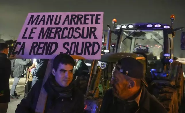 A farmer holds a placard that reads, "Macron stop the Mercosur it makes you deaf " as he stands next to tractors on a blocked highway in Velizy-Villacoublay, outside Paris, Sunday, Nov. 17, 2024. (AP Photo/Michel Euler)