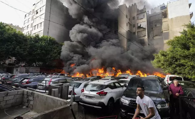 A man runs as a civil defense worker tries to extinguish a fire caused by a giant diesel tank which ignited, burning more than a dozen cars in a parking lot and damaging a building nearby, in a residential Beirut neighborhood, Lebanon, Saturday, Nov. 9, 2024. (AP Photo/Hassan Ammar)