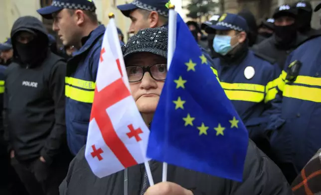 A woman with a EU and a Georgian national flags stands in front of police blocking a street during a rally to demand new parliamentary elections in the country, near the Parliament's building in Tbilisi, Georgia, on Monday, Nov. 25, 2024. (AP Photo/Shakh Aivazov)