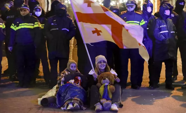 A woman and a girl sit holding a Georgian national flag in front of police blocking the entrance of the Parliament's building during a rally to demand new parliamentary elections in the country, in Tbilisi, Georgia, on Monday, Nov. 25, 2024. (AP Photo/Zurab Tsertsvadze)