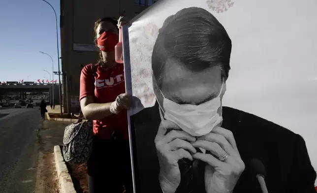 FILE - A demonstrator holds a banner showing Brazilian President Jair Bolsonaro adjusting his mask in Brasilia, Brazil, Aug. 7, 2020, as the Central Workers Unions (CUT) protest his handling of the new coronavirus pandemic. (AP Photo/Eraldo Peres, File)