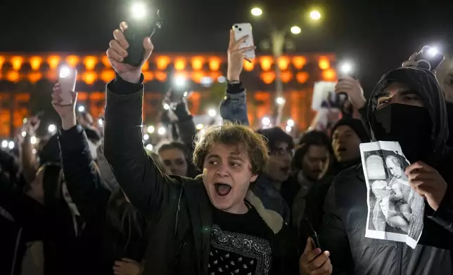 Youngsters shout slogans and flash the light of their mobile phones in Bucharest, Romania, Wednesday, Nov. 27, 2024, next to an altered version of a classic painting, depicting Russian President Vladimir Putin and Calin Georgescu, the independent candidate for Romanian presidency who won the first round of elections making it to the Dec. 8, runoff. (AP Photo/Vadim Ghirda)