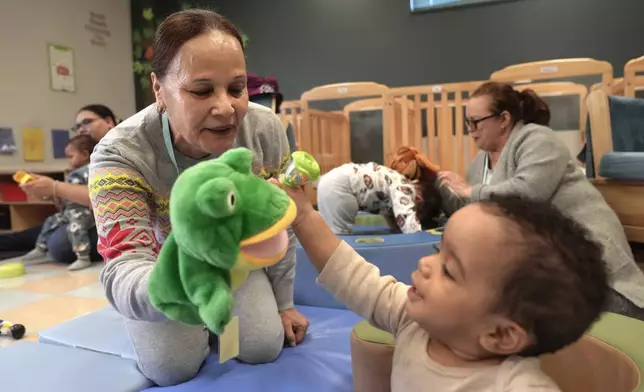 Volunteer Ydalina Luna Sosa, 62, of Providence, R.I., front left, uses a frog puppet for entertainment while supervising 10-month-old Leo Morgenweck, front right, in an early childcare program at Federal Hill House, Tuesday, Nov. 12, 2024, in Providence, R.I. (AP Photo/Steven Senne)