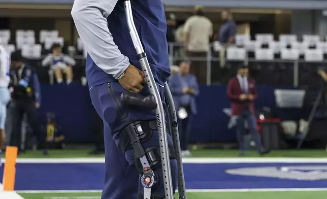 Dallas Cowboys quarterback Dak Prescott stands with crutches on the sidelines as teammates work out prior to an NFL football game against the Houston Texans, Monday, Nov. 18, 2024, in Arlington. (AP Photo/Gareth Patterson)