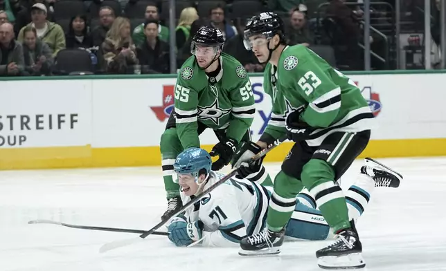 San Jose Sharks center Macklin Celebrini, center, falls to the ice after makiung a pass to the front of the net as Dallas Stars' Thomas Harley (55) and Wyatt Johnston (53) defend in the second period of an NHL hockey game in Dallas, Wednesday, Nov. 20, 2024. (AP Photo/Tony Gutierrez)