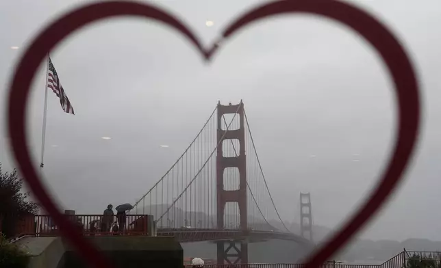 People carrying umbrellas while visiting the Golden Gate Bridge are seen through a heart on the window at the Round House Cafe in San Francisco, Wednesday, Nov. 20, 2024. (AP Photo/Jeff Chiu)