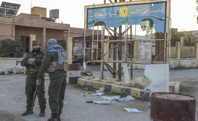 Syrian opposition fighters stand next to a government sign after entering the village of Anjara, western outskirts of Aleppo, Syria, Thursday Nov. 28, 2024, part of their major offensive on government-controlled areas in the country's northwestern Syria. (AP Photo/Omar Albam)