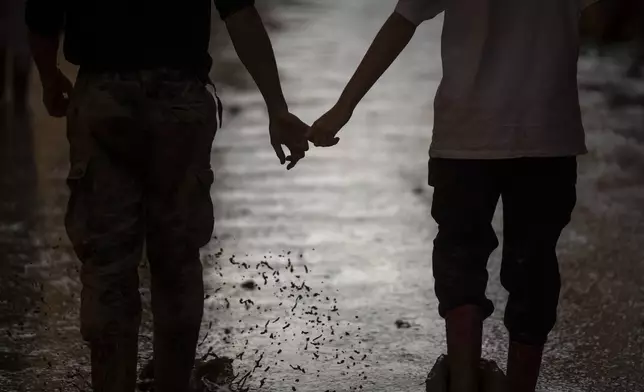 A couple of volunteers walk hand in hand down a muddy street in a flooded area in Masanasa, Valencia, Spain, Thursday, Nov. 7, 2024. (AP Photo/Emilio Morenatti)