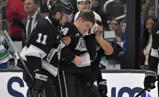 Los Angeles Kings defenseman Mikey Anderson, center, is helped off the ice by Los Angeles Kings center Anze Kopitar (11) after he was hit in the head with the puck during the second period of an NHL hockey game against the Vancouver Canucks, Thursday, Nov. 7, 2024, in Los Angeles. (AP Photo/Jayne-Kamin-Oncea)