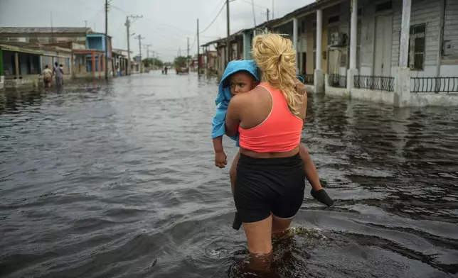 FILE - A woman carries a child as she wades through a street flooded in the passing of Hurricane Helene, in Batabano, Mayabeque province, Cuba, Sept. 26, 2024. (AP Photo/Ramon Espinosa, File)