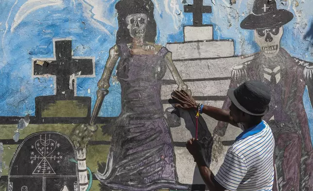 A Vodou believer touches a mural featuring calaveras during the annual Fete Gede festival that celebrates Day of the Dead, honoring the Haitian spirits Baron Samedi and Gede, at the National Cemetery, in Port-au-Prince, Haiti, Friday, Nov. 1, 2024. (AP Photo/Odelyn Joseph)