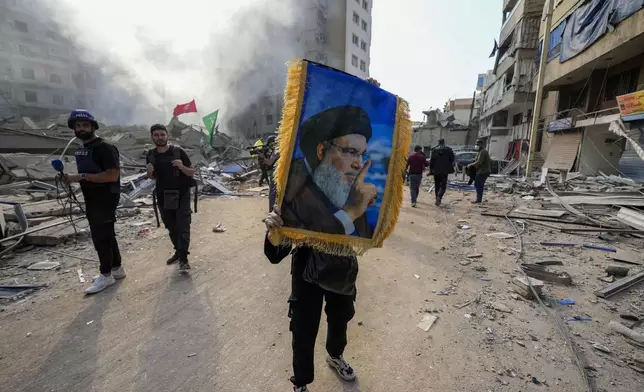 A woman holds a picture of slain Hezbollah leader Hassan Nasrallah as smoke rises from the site of an Israeli airstrike in Dahiyeh, Beirut, Lebanon, Friday, Nov. 1, 2024. (AP Photo/Hassan Ammar)