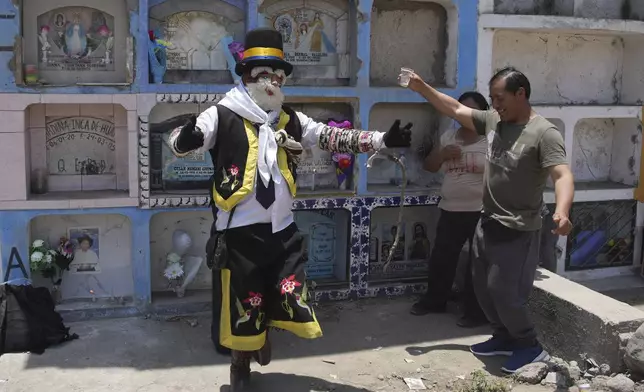 A performer dressed as the Andean character Chuto dances with Oswaldo Bellido who is visiting his parents' grave, during the Day of the Dead celebrations in Lima, Peru, Friday, Nov. 1, 2024, (AP Photo/Guadalupe Pardo)