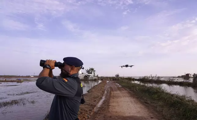 As the search for bodies continues, a Civil Guard looks though binoculars as a drone flies nearby at the mouth of the Poyo ravine in the La Albufera natural lake near Puerto de Catarroja, Valencia on the outskirts of Valencia, Spain, Tuesday, Nov. 5, 2024. (AP Photo/Alberto Saiz)