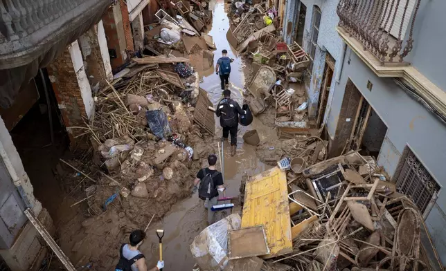 People walk through a street with piled furniture and rubbish on the sides in an area, affected by floods, in Paiporta, Valencia, Spain, Tuesday, Nov. 5, 2024. (AP Photo/Emilio Morenatti)