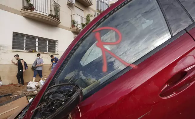 Volunteers helping with the clean up operation walk by a car with the letter R painted on the window meaning it has been revised as rescue workers look for bodies after floods in Massanassa, just outside of Valencia, Spain, Saturday, Nov. 2, 2024. (AP Photo/Alberto Saiz)