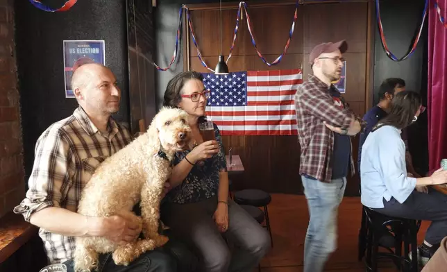 Steve Baker, left, Claudine Earley and their dog Louis watch a television broadcast during a U.S. election viewing party at Mean Doses bar in Wellington, New Zealand on Wednesday, Nov. 6, 2024. (AP Photo/Charlotte Graham-McLay)