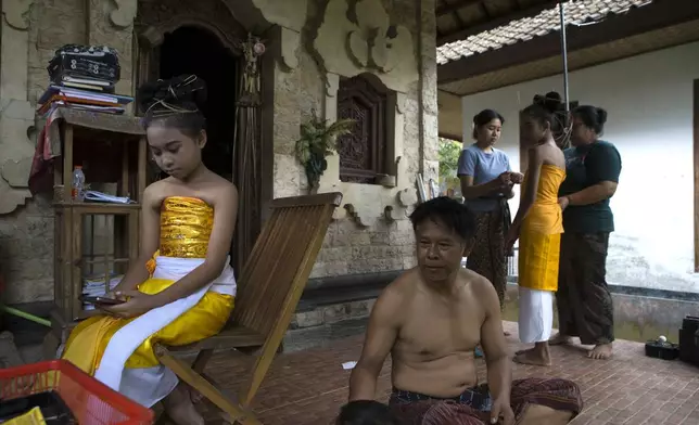 Ketut Nita Wahyuni, 11, looks at her phone with her father Nyoman Subrata sitting beside her, as mother Kadek Krisni, right, and sister Intan Wahyuni, in blue, dresses up her cousin Rina Lestianti before the Rejang Pucuk Hindu rituals at Geriana Kauh village, Karangasem, Bali, Indonesia on Thursday, Nov. 21, 2024. (AP Photo/Firdia Lisnawati)