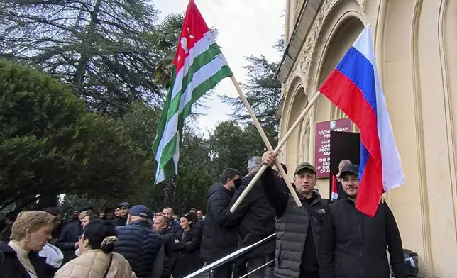 In this photo taken from video released by AIASHARA Independent Agency, Protesters, two of them hold an Abkhazian and Russian flags, gather outside the parliament building of the Georgian separatist region of Abkhazia as tensions flared over a proposed pact that would allow Russians to buy apartments in the region, Georgia, on Friday, Nov. 15, 2024, (AIASHARA Independent Agency via AP)