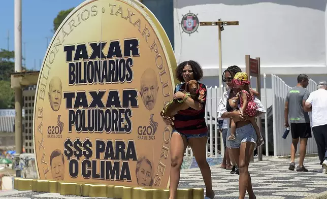 Women walk past a giant coin that reads in Portuguese "Tax billionaires, tax polluters, $$$ for climate" on Leblon beach as part of a protest to draw attention to climate issues on the sidelines of the G20 Summit in Rio de Janeiro, Monday, Nov. 18, 2024. (AP Photo/Dhavid Normando)