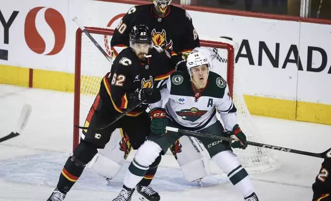 Minnesota Wild's Joel Eriksson Ek (14) is checked by Calgary Flames' Daniil Miromanov (16) as Flames goalie Dustin Wolf (80) looks on during first-period NHL hockey game action in Calgary, Alberta, Saturday, Nov. 23, 2024. (Jeff McIntosh/The Canadian Press via AP)