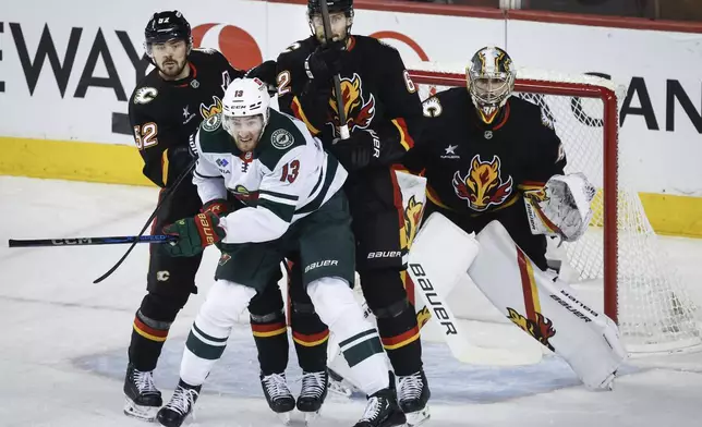 Minnesota Wild's Yakov Trenin (13) is checked by Calgary Flames MacKenzie Weegar (52) and Daniil Miromanov (62) as Flames goalie Dustin Wolf, right, looks on during first-period NHL hockey game action in Calgary, Alberta, Saturday, Nov. 23, 2024. (Jeff McIntosh/The Canadian Press via AP)