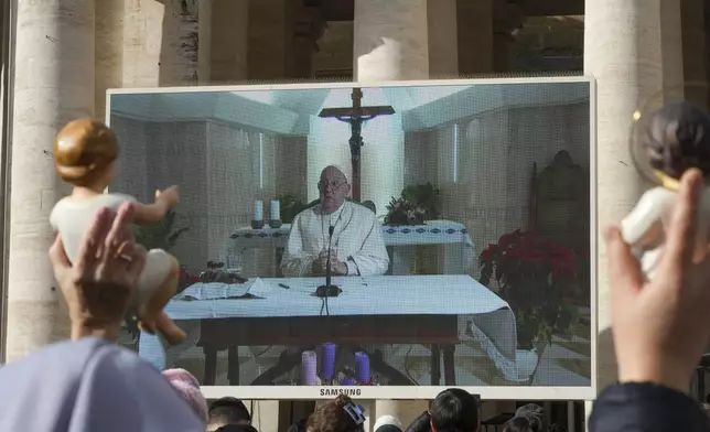 People hold statuettes of baby Jesus as they look at a giant screen in St.Peter's Square, at the Vatican, showing Pope Francis reciting the Angelus noon prayer from his Santa Marta Residence, after he decided not to appear at the window of his studio overlooking the square because of a persistent cold, Sunday, Dec. 22, 2024. (AP Photo/Andrew Medichini)