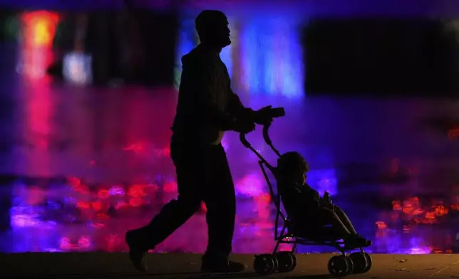 A person pushes a child in a stroller as they are silhouetted against holiday lights reflected on a pond in a park, Saturday, Dec. 7, 2024, in Lenexa, Kan. (AP Photo/Charlie Riedel)