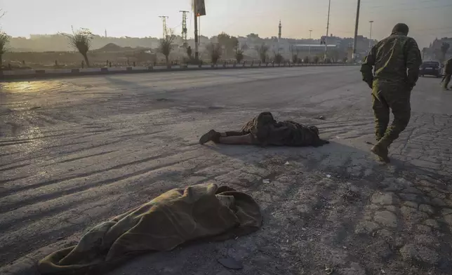 An opposition fighter walks past the bodies of two government soldiers lying on the ground in the aftermath of the opposition's takeover in Hama, Syria, Friday, Dec. 6, 2024. (AP Photo/Omar Albam)